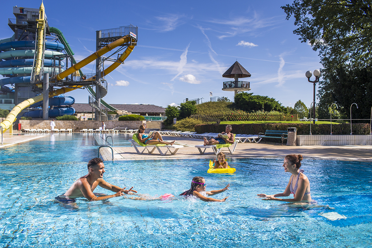 Mlajša deklica se v zunanjem bazenu uči plavati, oče in mama pa ji pri tem pomagata