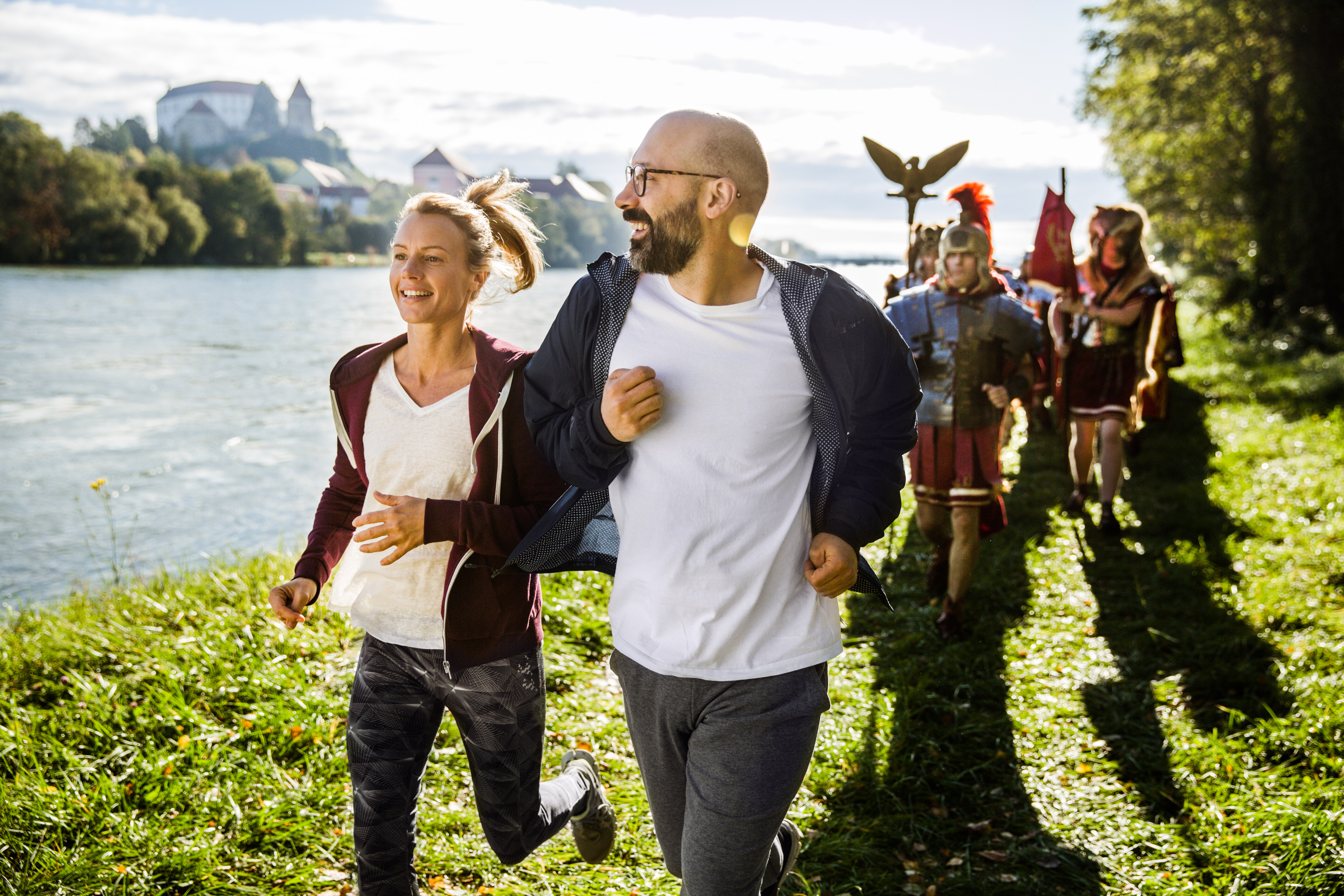 Moški in ženska v športnih oblačilih tečeta ob reki pred skupino ljudi, oblečenih v srednjeveške viteze