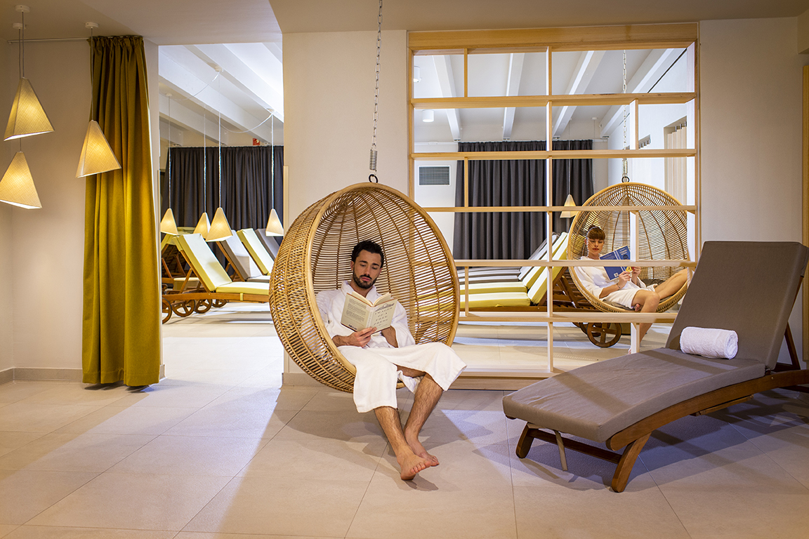 Moški v belem kopalnem plašču sedi na visečem sedežu ob ležalniku in bere knjigo